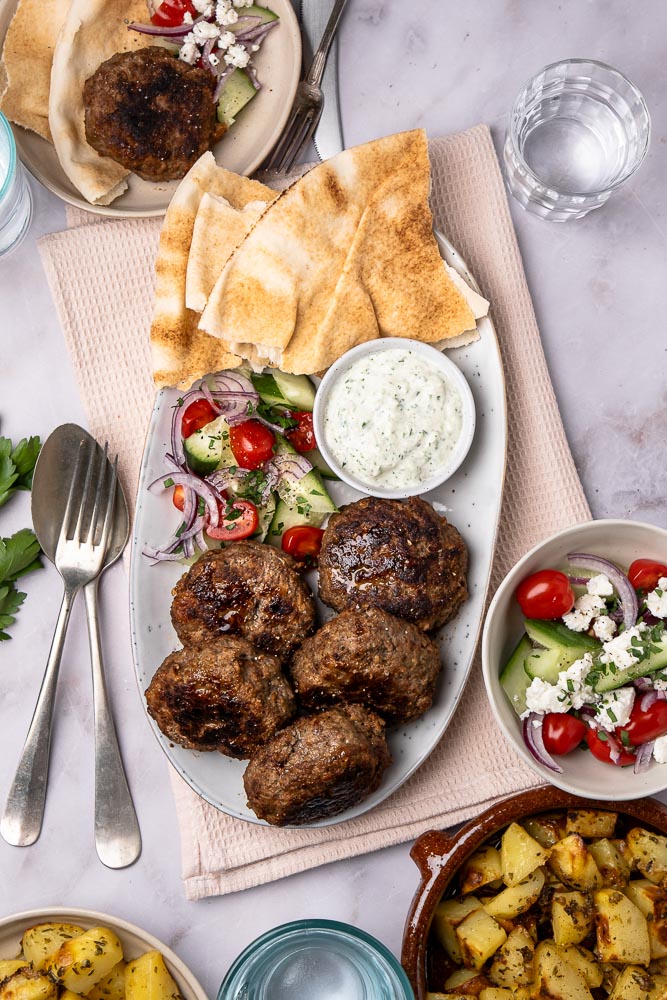 Traditionele Griekse bifteki gehaktballen recept, gerecht met gehakt, tomaat, ui en verse kruiden en geserveerd met tzatziki saus en een frisse Griekse salade.