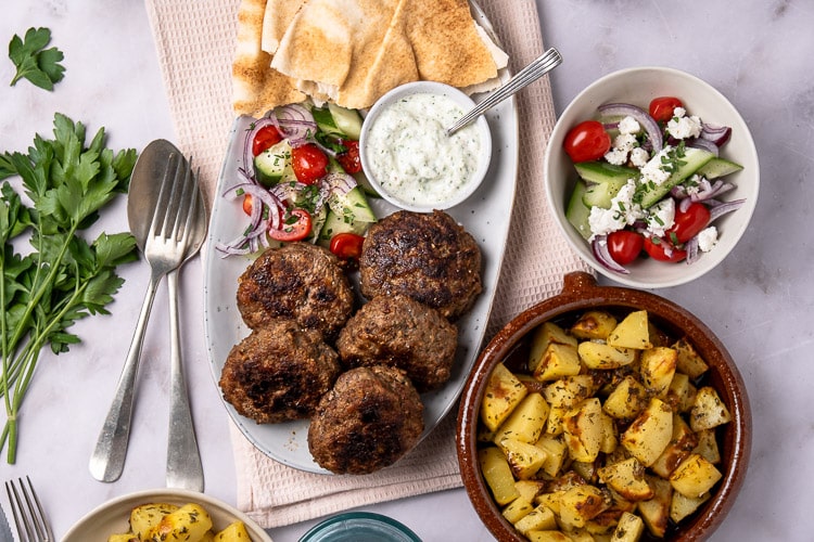 Traditionele Griekse bifteki gehaktballen recept, gerecht met gehakt, tomaat, ui en verse kruiden en geserveerd met tzatziki saus en een frisse Griekse salade.