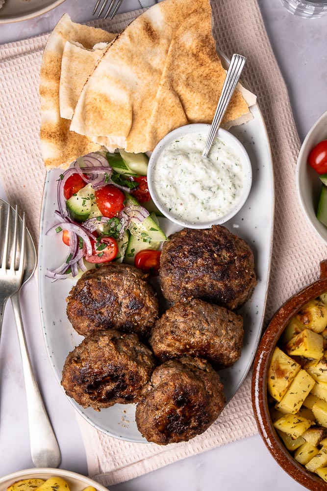 Traditionele Griekse bifteki gehaktballen recept, gerecht met gehakt, tomaat, ui en verse kruiden en geserveerd met tzatziki saus en een frisse Griekse salade.