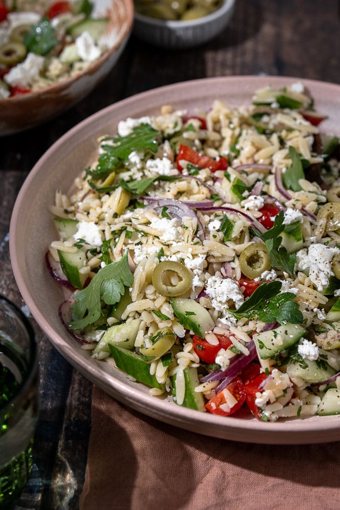 Serveersuggestie van Griekse orzo salade voor de zomer. Ontdek de heerlijke smaken van een Griekse orzo salade, een frisse en gezonde keuze voor elke maaltijd. Perfect als bijgerecht of hoofdgerecht met ingrediënten zoals feta, olijven, tomaat en komkommer. Ideaal voor zomerse dagen!