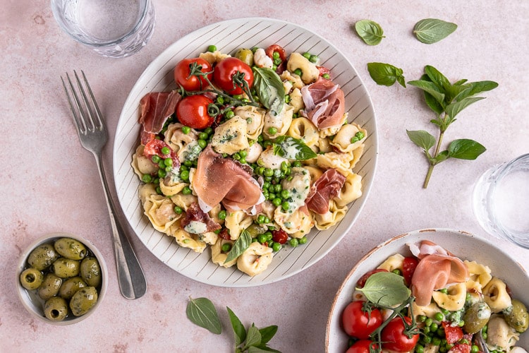 Italiaanse pastasalade met prosciutto, mozzarella, tomaatjes, olijven, pancetta en doperwtjes, geserveerd met een frisse balsamico dressing en verse basilicum.