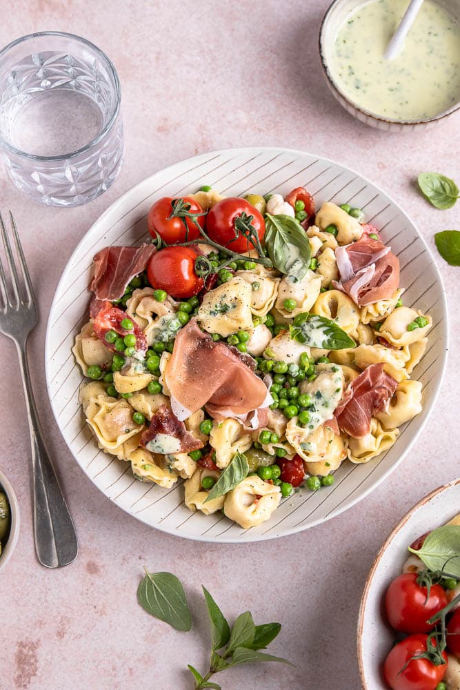 taliaanse pastasalade met prosciutto, mozzarella, tomaatjes, olijven, pancetta en doperwtjes, geserveerd met een frisse balsamico dressing en verse basilicum.