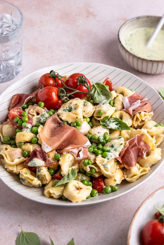 taliaanse pastasalade met prosciutto, mozzarella, tomaatjes, olijven, pancetta en doperwtjes, geserveerd met een frisse balsamico dressing en verse basilicum.