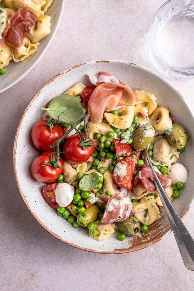 taliaanse pastasalade met prosciutto, mozzarella, tomaatjes, olijven, pancetta en doperwtjes, geserveerd met een frisse balsamico dressing en verse basilicum.