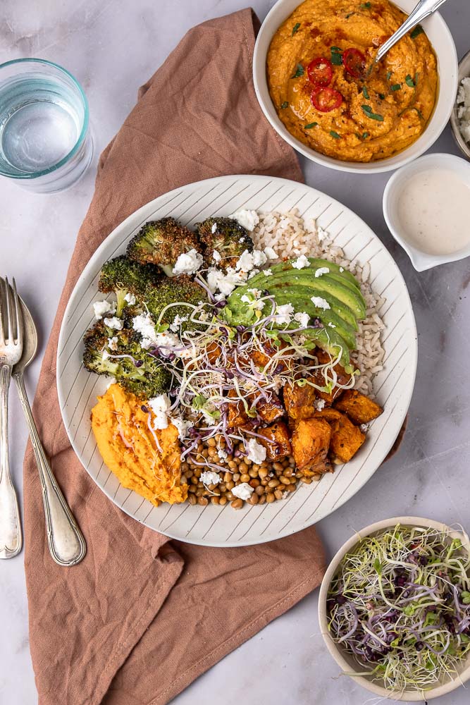 Makkelijk recept voor buddha bowl met geroosterde zoete aardappel, broccoli, volkoren rijst, linzen, feta, avocado, hummus met geroosterde paprika en groentekiemen, geserveerd in een kom. Een gezonde, vegetarische maaltijd vol voedzame ingrediënten.