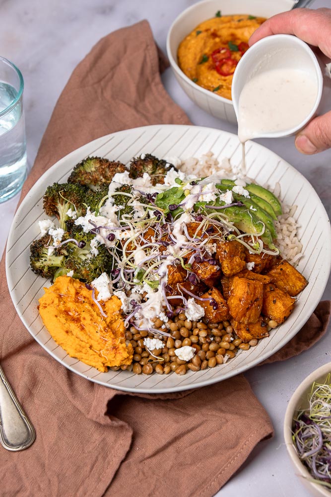 Makkelijk recept voor buddha bowl met geroosterde zoete aardappel, broccoli, volkoren rijst, linzen, feta, avocado, hummus met geroosterde paprika en groentekiemen, geserveerd in een kom. Een gezonde, vegetarische maaltijd vol voedzame ingrediënten.