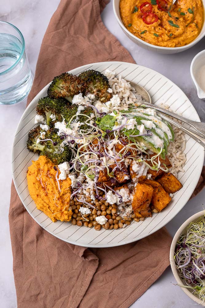 Makkelijk recept voor buddha bowl met geroosterde zoete aardappel, broccoli, volkoren rijst, linzen, feta, avocado, hummus met geroosterde paprika en groentekiemen, geserveerd in een kom. Een gezonde, vegetarische maaltijd vol voedzame ingrediënten.