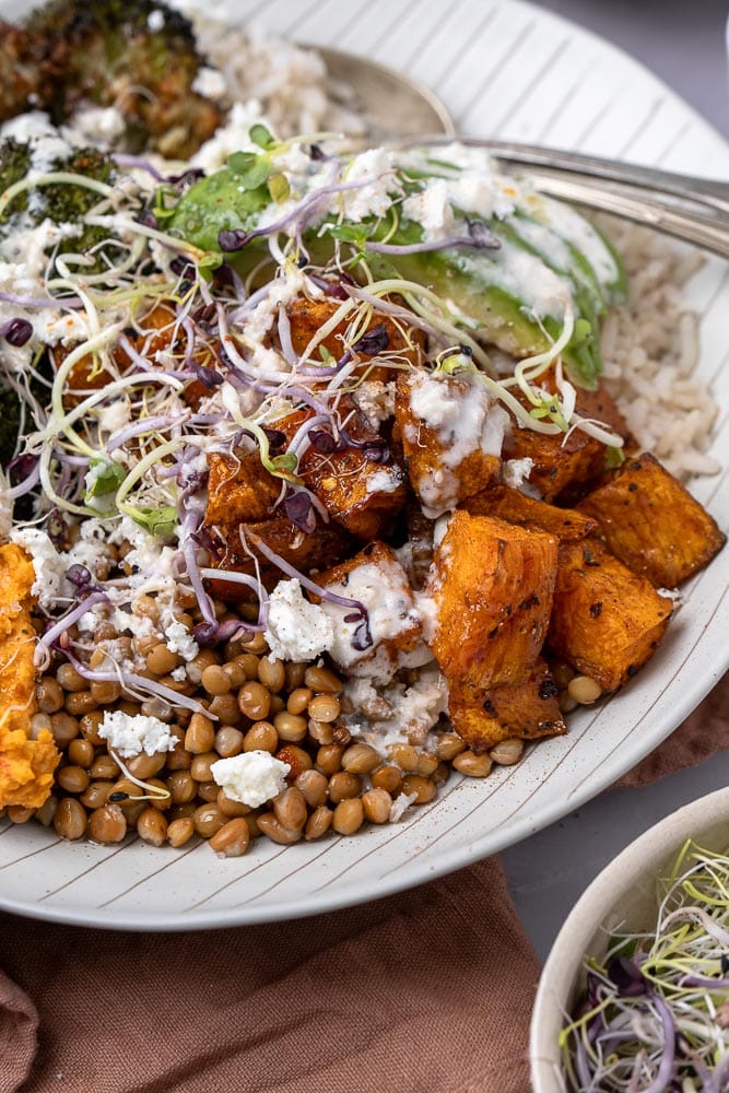 Makkelijk recept voor buddha bowl met geroosterde zoete aardappel, broccoli, volkoren rijst, linzen, feta, avocado, hummus met geroosterde paprika en groentekiemen, geserveerd in een kom. Een gezonde, vegetarische maaltijd vol voedzame ingrediënten.