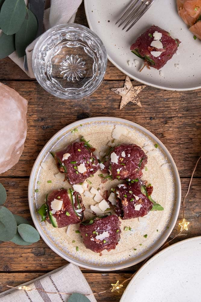 Carpaccio rolletjes met komkommer, truffelmayonaise, krokante spekkruimels, Parmezaanse kaas, zongedroogde tomaatjes, rucola en bieslook, geserveerd als feestelijk voorgerecht. Carpaccio hapje als amuse voor een Kerst diner, carpaccio koolhydraatarm voorgerecht.