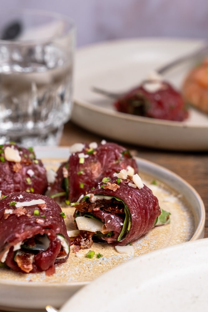 Carpaccio rolletjes met komkommer, truffelmayonaise, krokante spekkruimels, Parmezaanse kaas, zongedroogde tomaatjes, rucola en bieslook, geserveerd als feestelijk voorgerecht. Carpaccio hapje als amuse voor een Kerst diner, carpaccio koolhydraatarm voorgerecht.