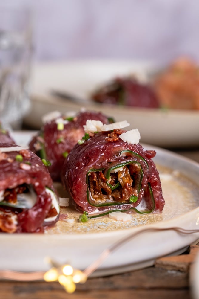 Carpaccio rolletjes met komkommer, truffelmayonaise, krokante spekkruimels, Parmezaanse kaas, zongedroogde tomaatjes, rucola en bieslook, geserveerd als feestelijk voorgerecht. Carpaccio hapje als amuse voor een Kerst diner, carpaccio koolhydraatarm voorgerecht.