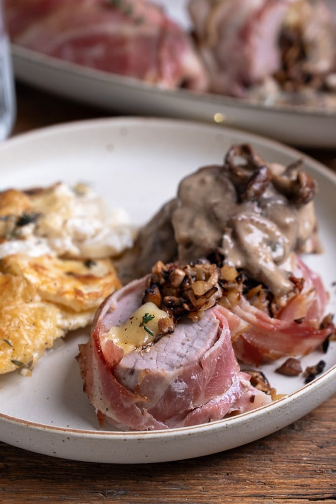 Gevulde varkenshaas met brie, champignons, zongedroogde tomaatjes en walnoten, omwikkeld met spek en gebakken in de oven, geserveerd als feestelijk hoofdgerecht. Recept voor gevulde varkenshaas met brie en spek met champignons en champignonroomsaus, feestelijk hoofgerecht met varkenshaas, bereidingswijze tips.